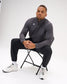 Man wearing the carbon RUDIS Victory 1/4 Zip with black athletic pants and white training shoes in a seated studio setting.