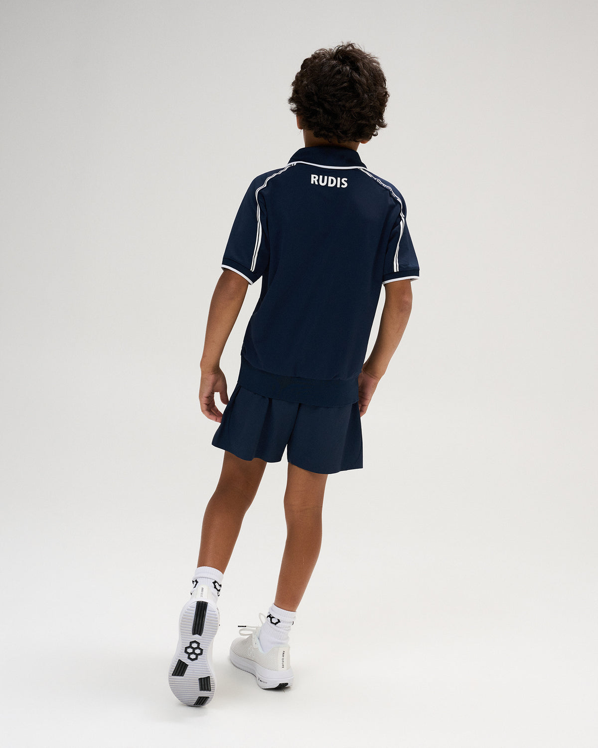 Back view of a youth model wearing the RUDIS Chronicle Elite Youth Athletic Polo in Navy, highlighting white RUDIS lettering below the collar, contrast piping along the shoulders, and short sleeves.