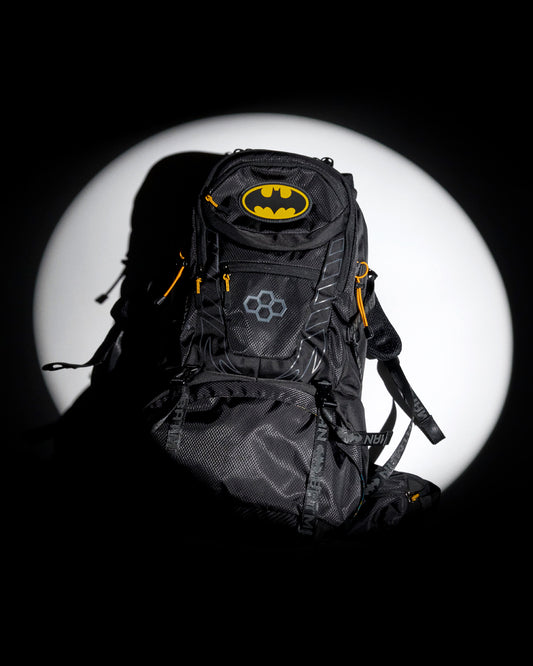 A black Batman-themed backpack with yellow and gray logo details, set against a dramatic black background with a large white spotlight circle behind it.