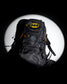 A black Batman-themed backpack with yellow and gray logo details, set against a dramatic black background with a large white spotlight circle behind it.