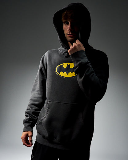 A man wearing a dark gray hooded sweatshirt with a yellow Batman logo on the chest, posed against a dark gray studio background.