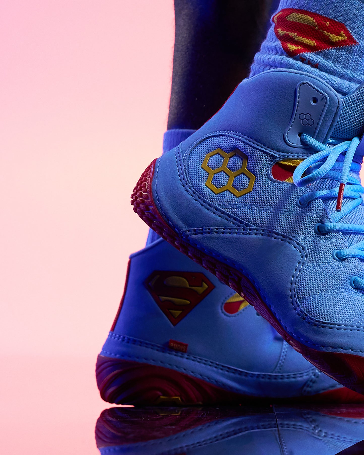 A close-up of a light blue high-top wrestling shoe with red and yellow Superman-style logos, shown on a foot against a pink studio background.