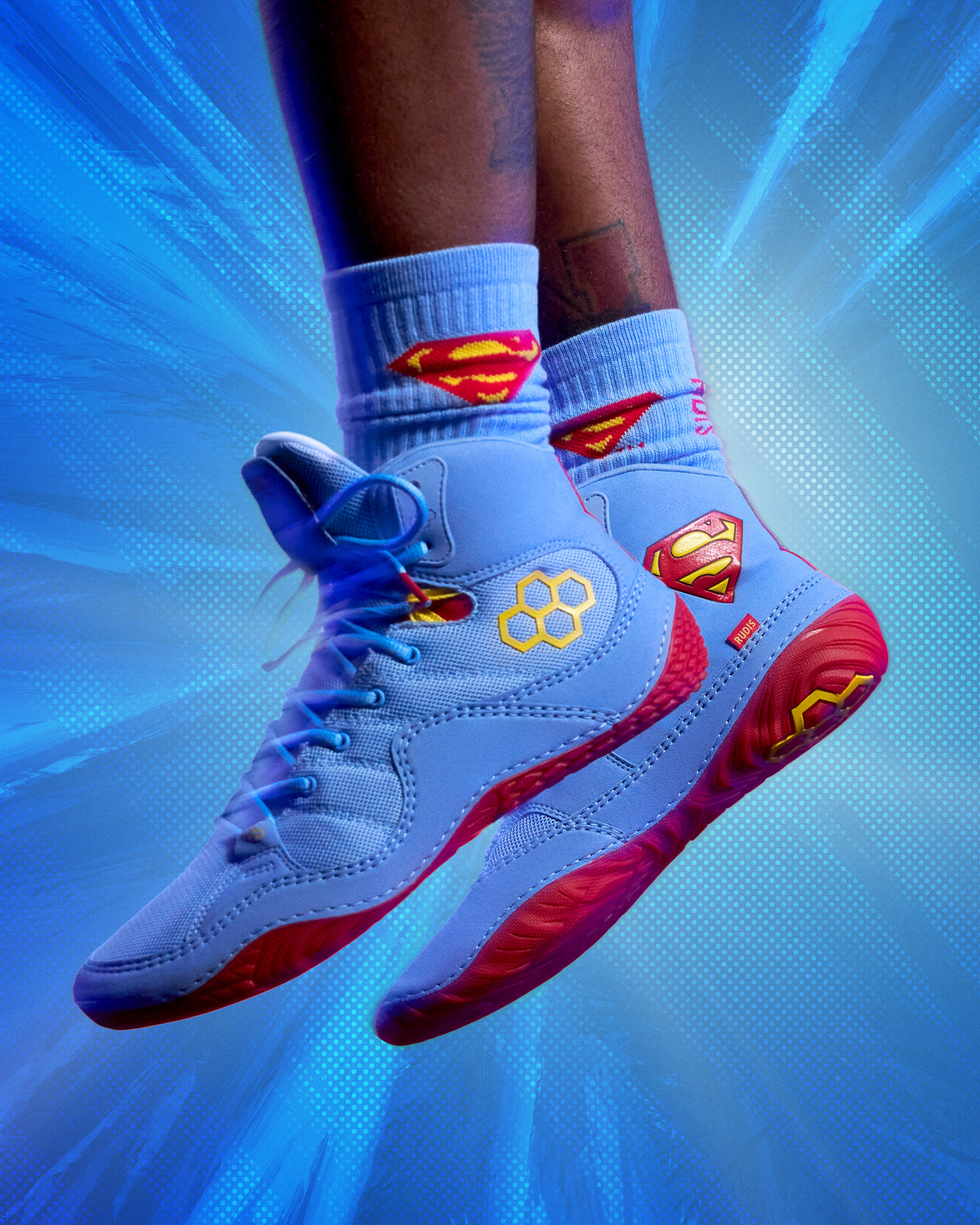 RUDIS JB1 Superman wrestling shoes worn with matching Superman socks against a comic-inspired blue and pink background.