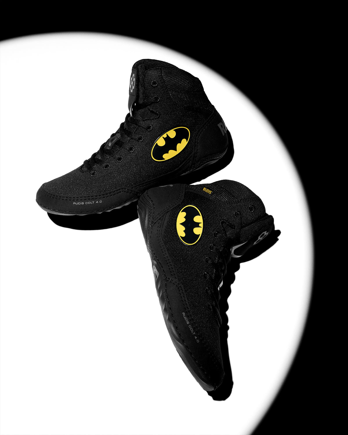 Two black high-top wrestling shoes with yellow Batman logos, floating against a white circular spotlight on a black background.