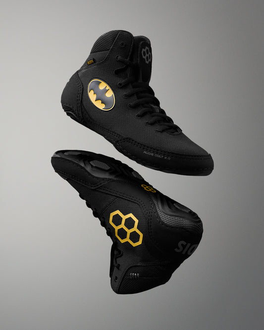 Black high-top wrestling shoes with gold Batman logo and honeycomb pattern accents, shown from multiple angles on a gray gradient background.
