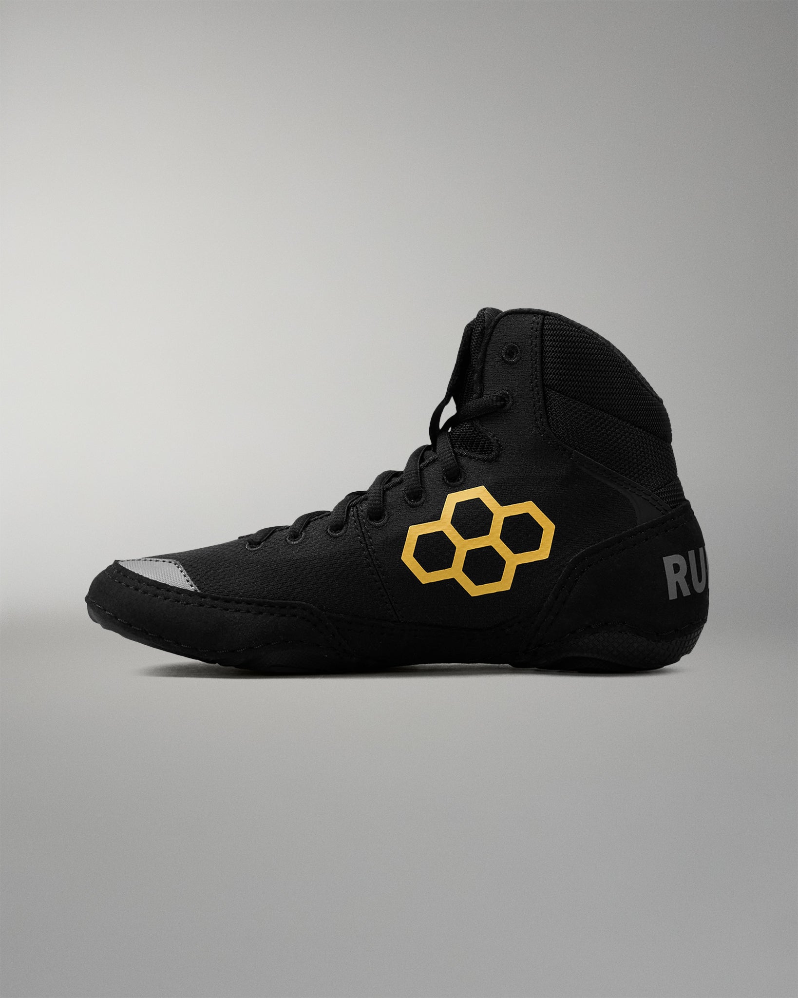 A black high-top wrestling shoe with a prominent yellow hexagonal logo on the side, a metallic silver toe cap, and partial white branding text on the heel, shown against a neutral gray background.