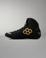 A black high-top wrestling shoe with a prominent yellow hexagonal logo on the side, a metallic silver toe cap, and partial white branding text on the heel, shown against a neutral gray background.