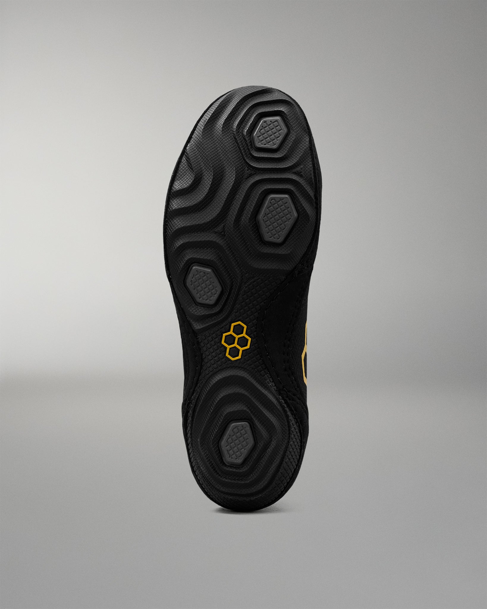 The black textured sole of a wrestling shoe, shown from a top-down view against a neutral gray background. The sole features a honeycomb-like tread pattern and a small yellow logo in the center.