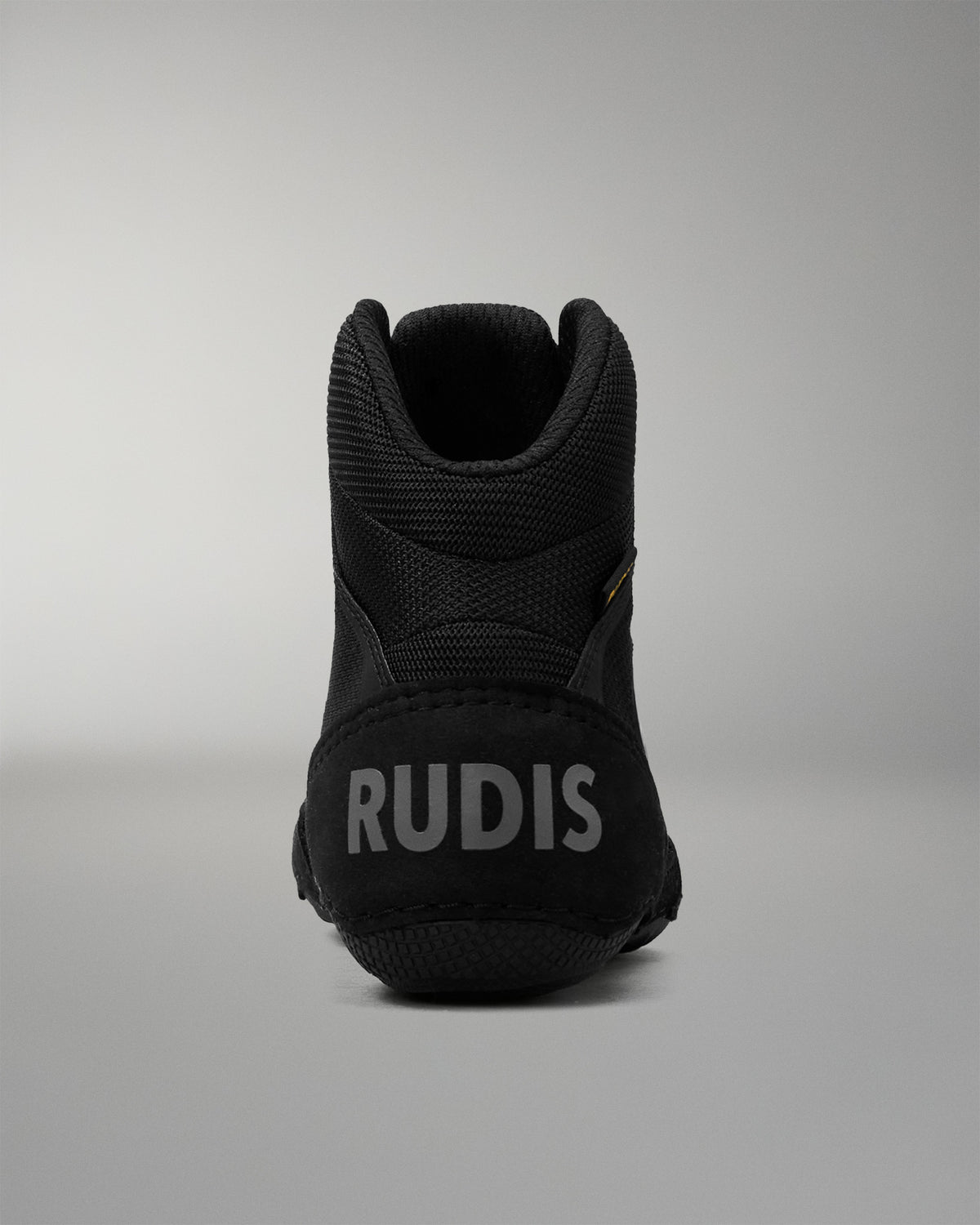 A black high-top wrestling shoe shown from the back, featuring the brand name 'RUDIS' in light gray text on the heel, set against a neutral gray background.