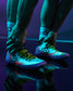 A close-up of a person's legs and hands adjusting colorful athletic wrestling shoes with matching patterned socks, set against a dark studio background with dramatic blue and purple lighting and a reflective floor.