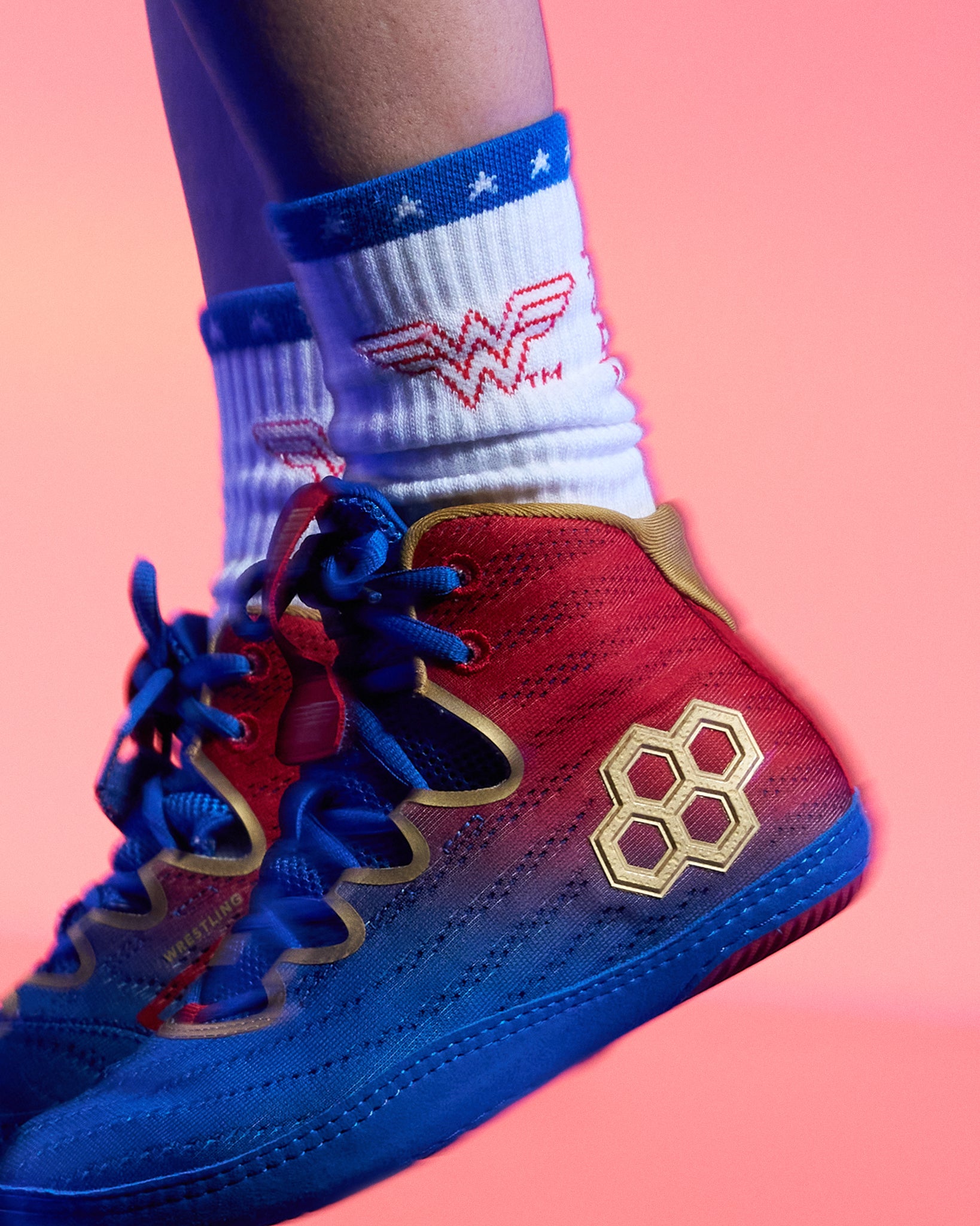 A close-up of a person's foot wearing a red, blue, and gold high-top wrestling shoes with a gold hexagon logo on the side, paired with white socks featuring a red and blue Wonder Woman logo, set against a solid pink background.