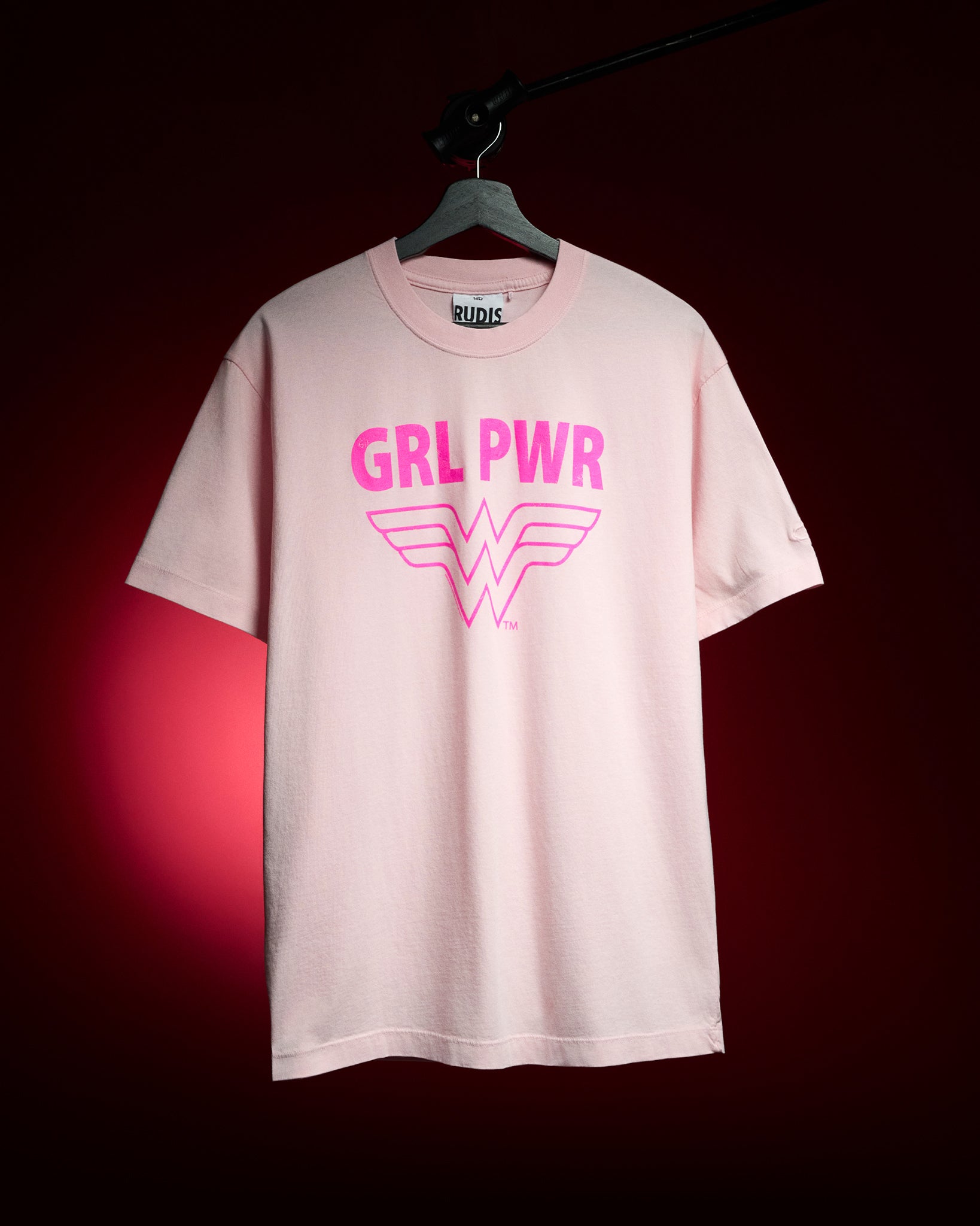 RUDIS Wonder Woman Girl Power Relaxed Cotton T-Shirt in washed pink, featuring a bold hot pink GRL PWR graphic and Wonder Woman logo, displayed on a hanger against a red background.