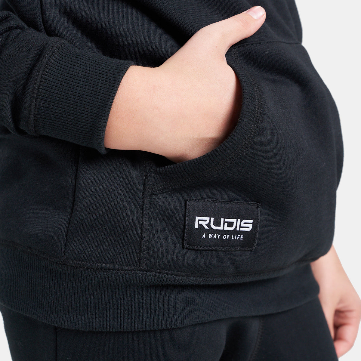A close-up view of a child wearing a black sweatshirt with a side pocket and a label that reads RUDIS A WAY OF LIFE