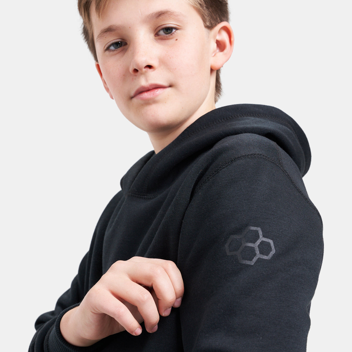 A young boy poses confidently in a black hoodie highlighting a unique logo on his left shoulder and a slight smile on his face