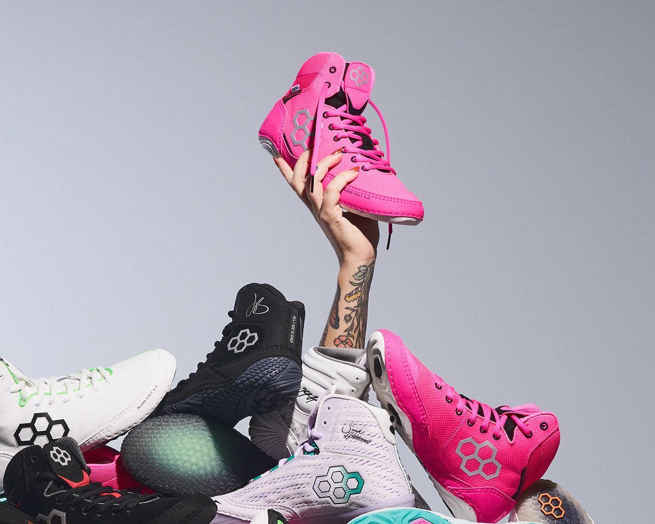 Hand holding up bright pink RUDIS wrestling shoes above a pile of RUDIS performance  and wrestling footwear, showcasing bold colorways, signature athlete designs, and premium wrestling shoe innovation.