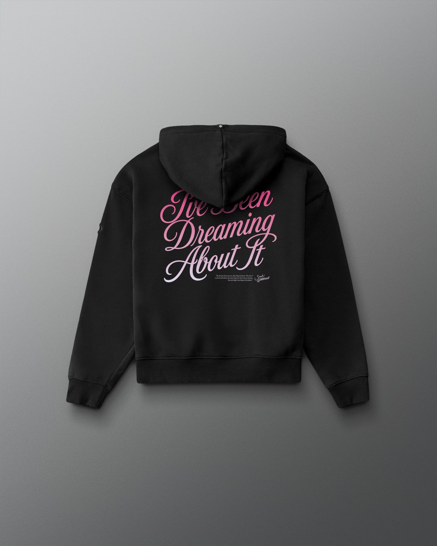 A black hoodie shown from the back, featuring the phrase 'I've Been Dreaming About It' printed in pink and white script across the upper back, displayed against a dark gray gradient background.