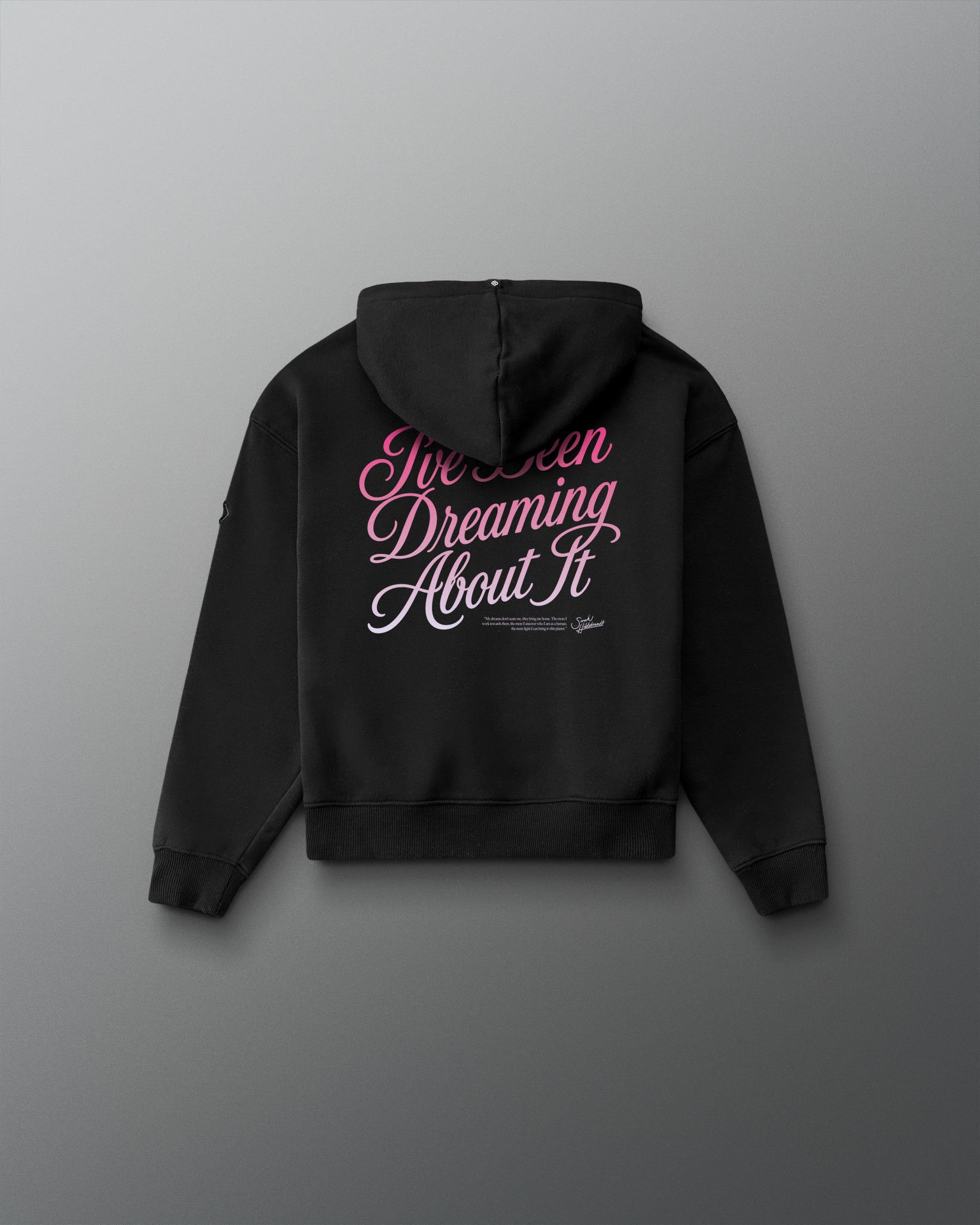 A black hoodie shown from the back, featuring the phrase 'I've Been Dreaming About It' printed in pink and white script across the upper back, displayed against a dark gray gradient background.