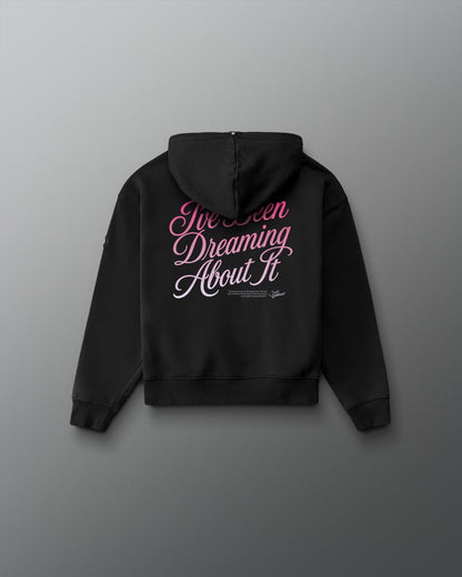 A black hoodie shown from the back, featuring the phrase 'I've Been Dreaming About It' printed in pink and white script across the upper back, displayed against a dark gray gradient background.