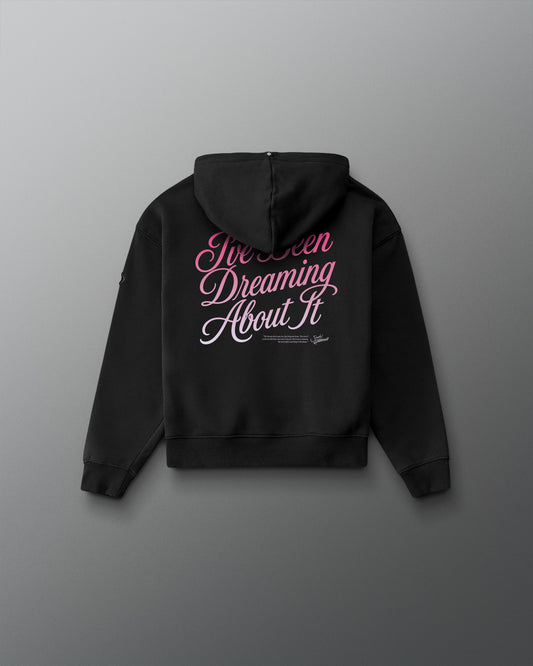 A black hoodie shown from the back, featuring the phrase 'I've Been Dreaming About It' printed in pink and white script across the upper back, displayed against a dark gray gradient background.