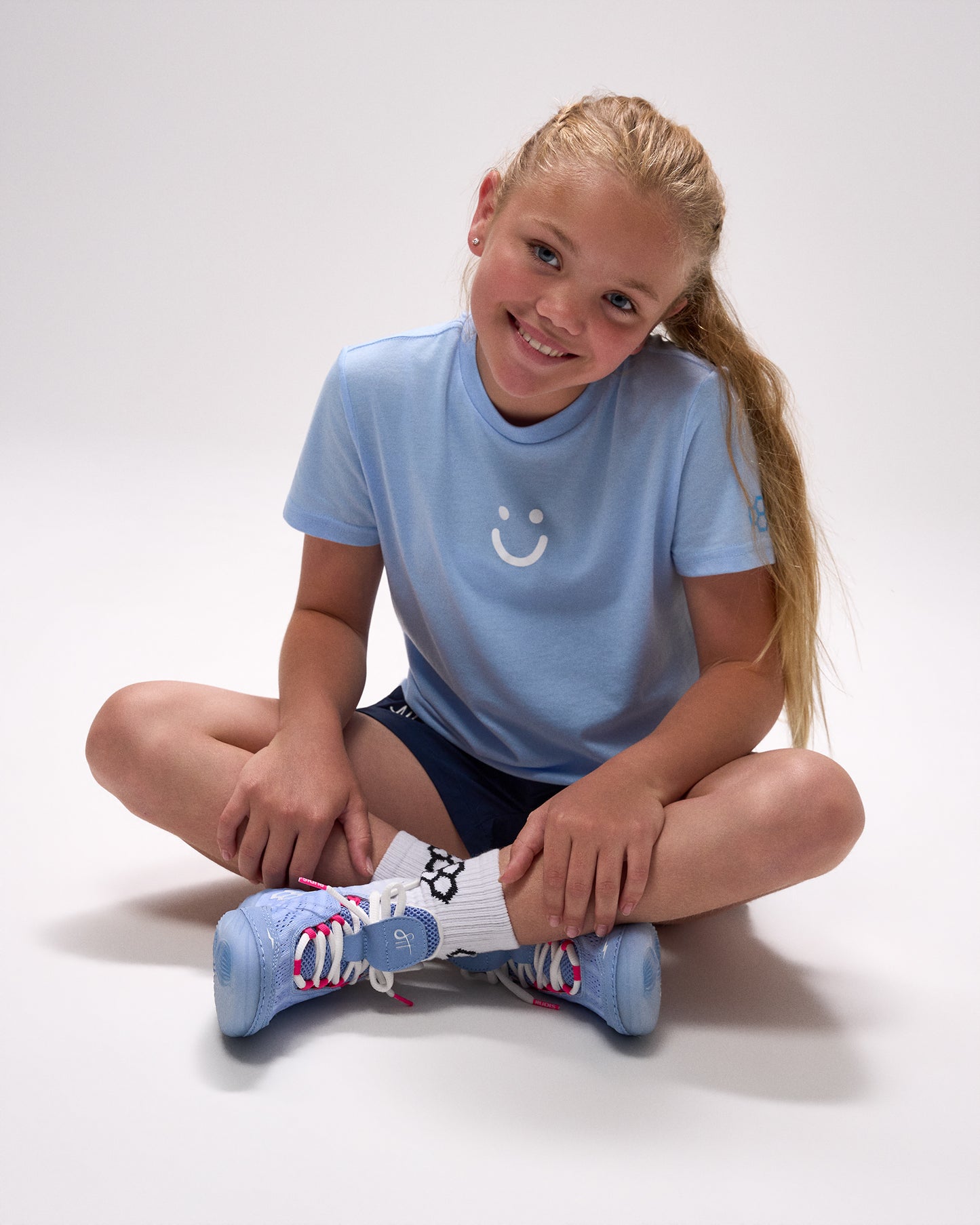 Young girl wearing a light blue t-shirt with a smiley face design, sitting on the floor with colorful socks.