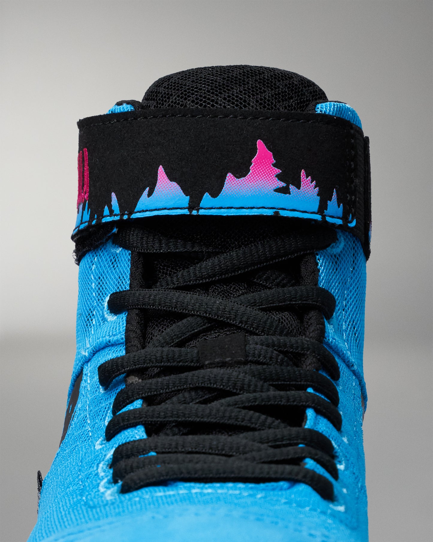 Close-up of a high-top wrestling shoe featuring a bright blue textured canvas upper, black laces, a black ankle strap, and a vibrant pink-to-blue gradient mountain silhouette graphic on the strap.