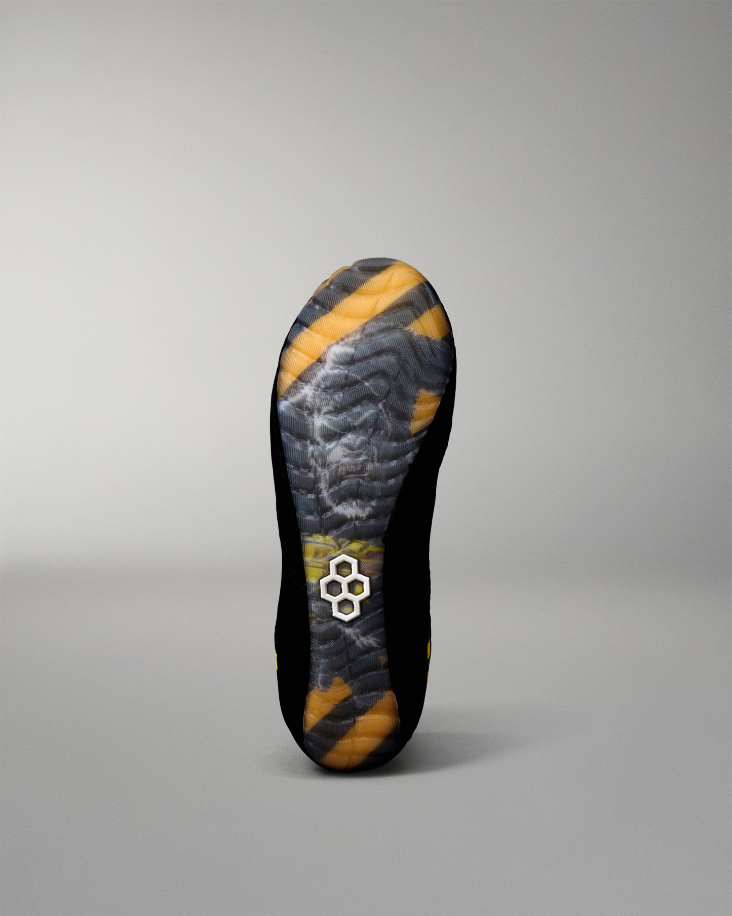 The outsole of a black wrestling shoe with yellow and gray patterned tread, featuring a small white geometric logo near the heel, shown against a neutral gray background.