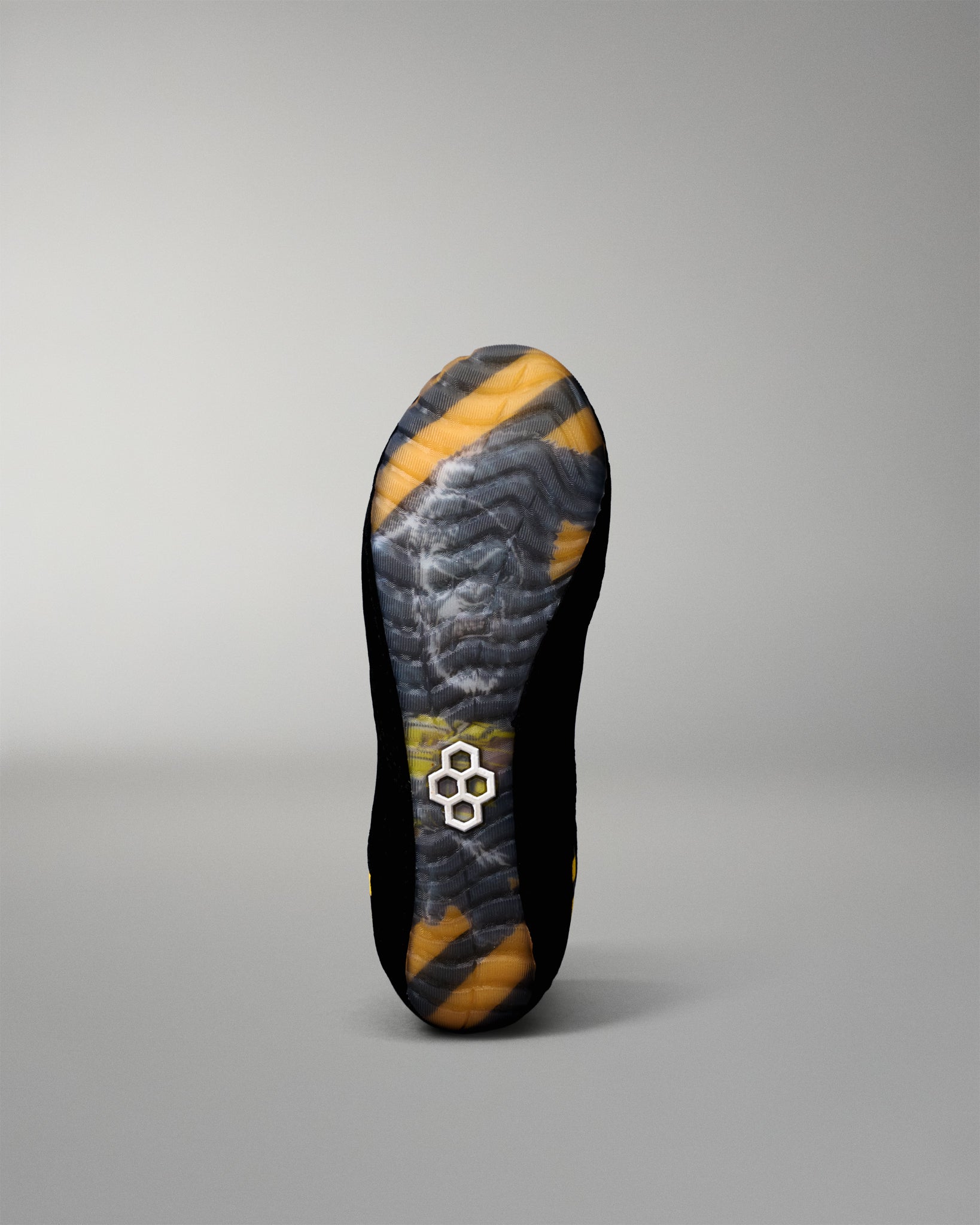 The outsole of a black wrestling shoe with yellow and gray patterned tread, featuring a small white geometric logo near the heel, shown against a neutral gray background.