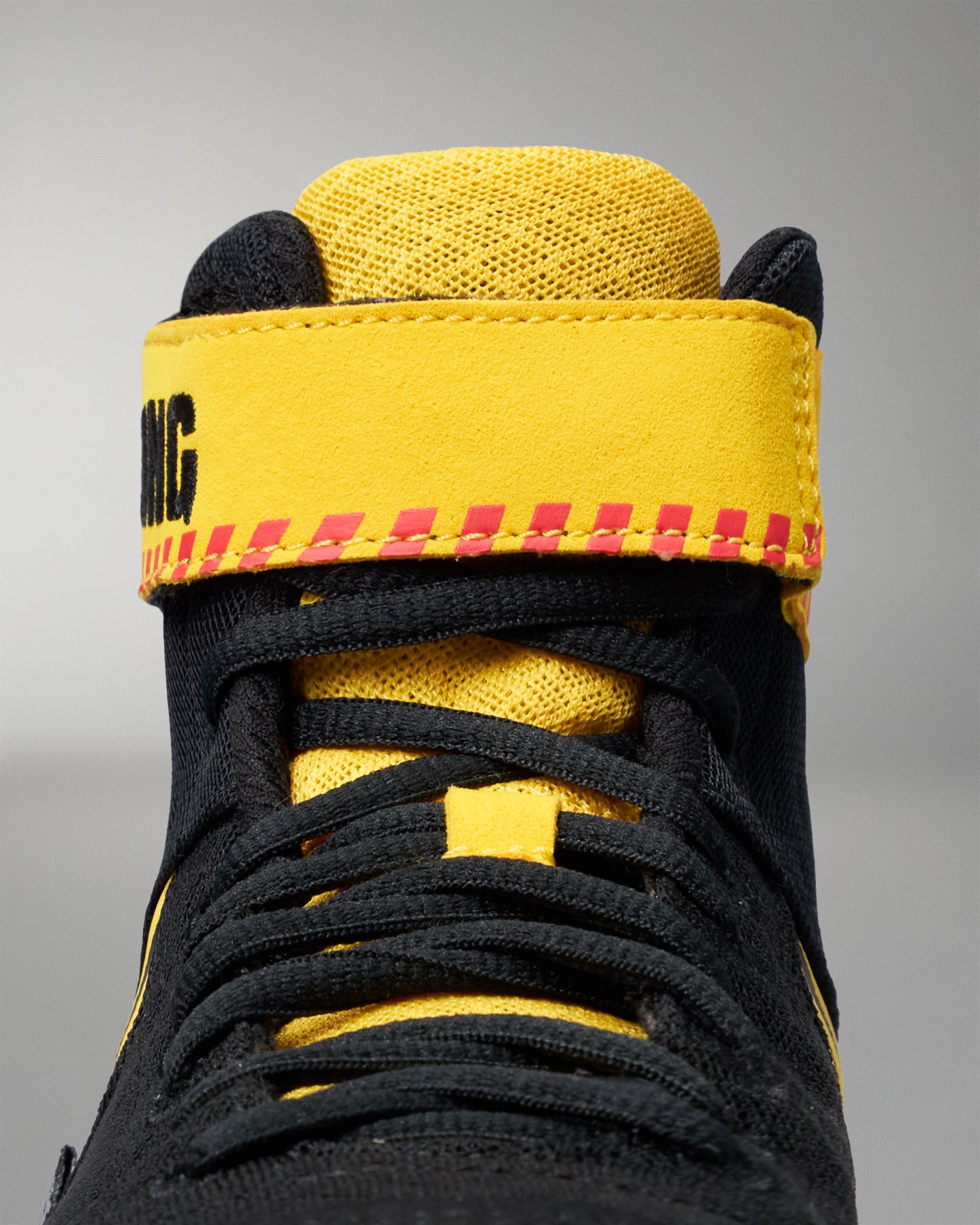 Close-up of a high-top wrestling shoe in black and bright yellow, showing the ankle strap with red stitching and black lettering, black laces, and a textured yellow collar on a gray background.