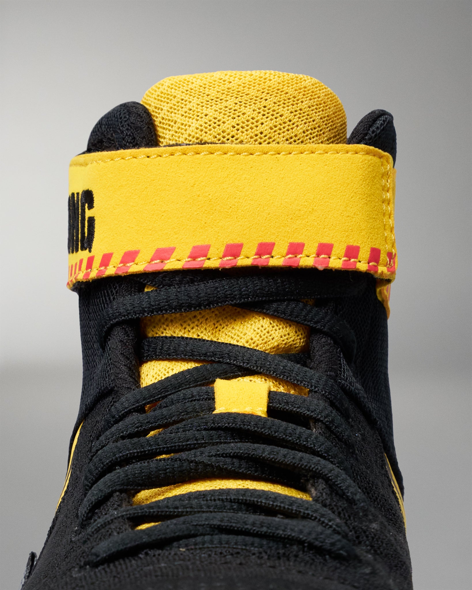 Close-up of a high-top wrestling shoe in black and bright yellow, showing the ankle strap with red stitching and black lettering, black laces, and a textured yellow collar on a gray background.