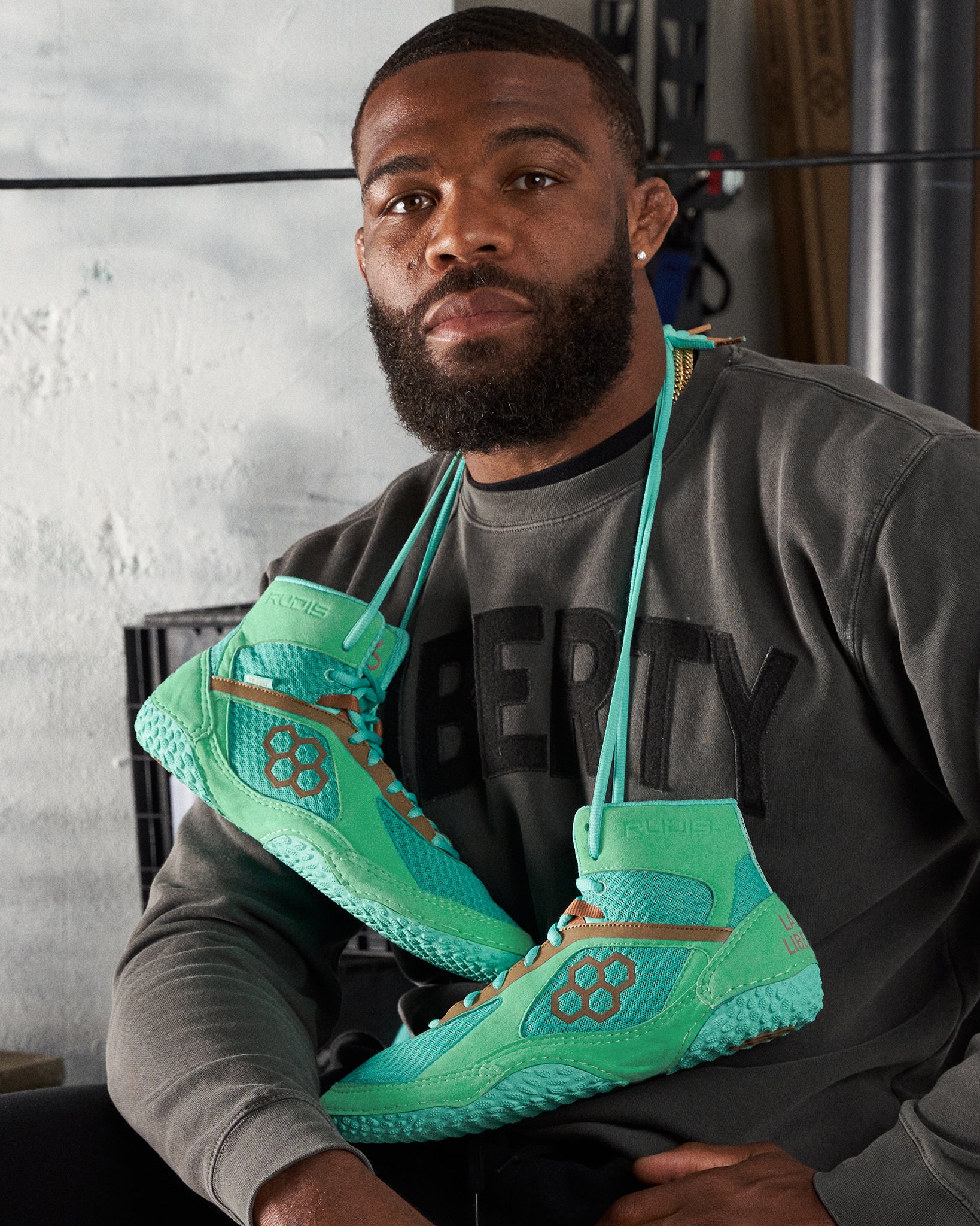 A man wearing a dark gray sweatshirt with LIBERTY printed on it poses while holding vibrant green athletic shoes that feature a unique design and textured soles highlighting their performance qualities