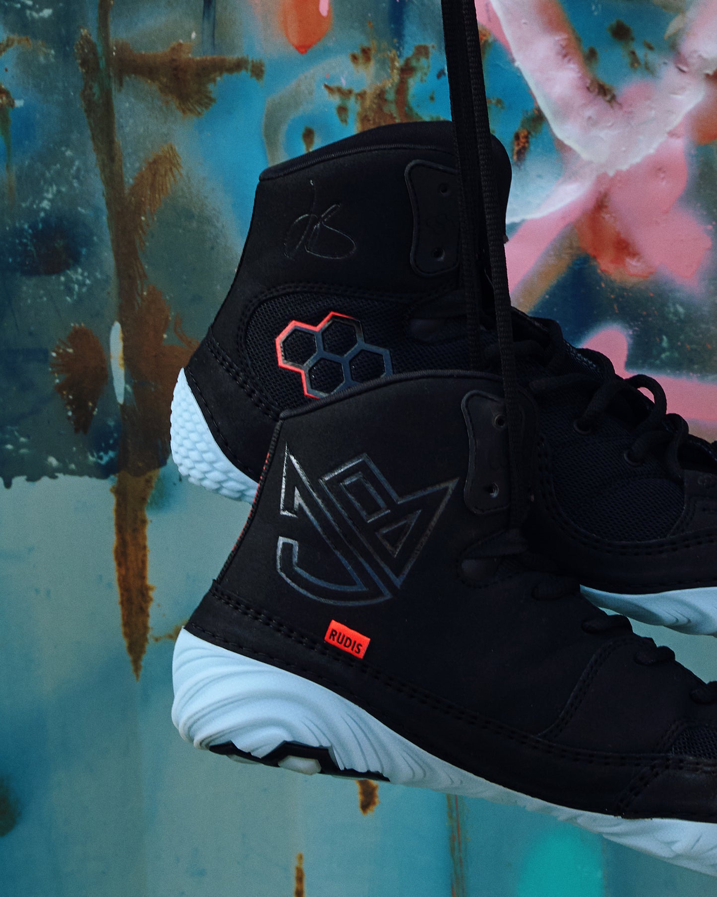 Youth RUDIS JB1 Dark Shark wrestling shoes in black with icy blue outsoles, JB signature branding, and red-accented RUDIS logo, suspended against an artistic graffiti backdrop.