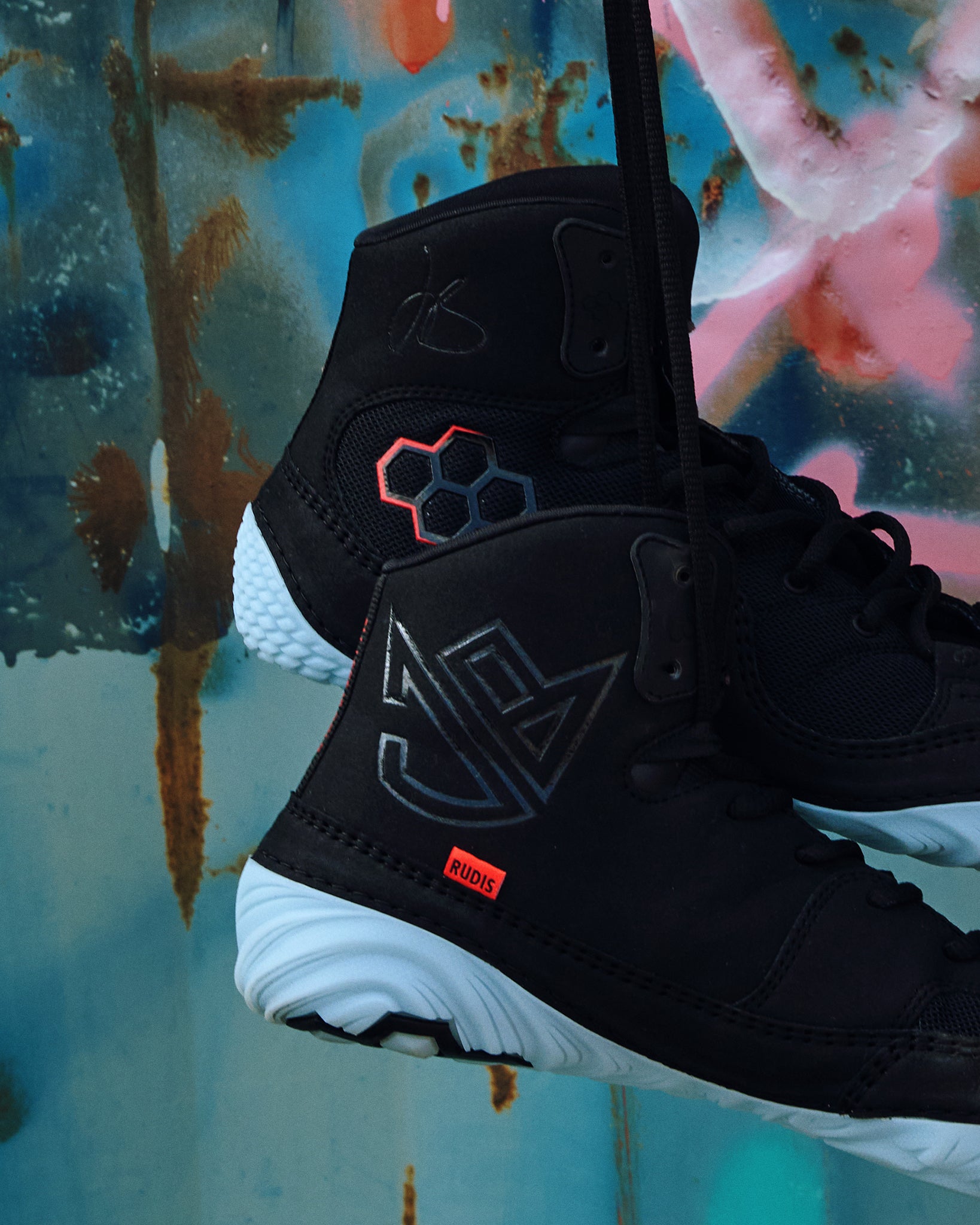 Youth RUDIS JB1 Dark Shark wrestling shoes in black with icy blue outsoles, JB signature branding, and red-accented RUDIS logo, suspended against an artistic graffiti backdrop.