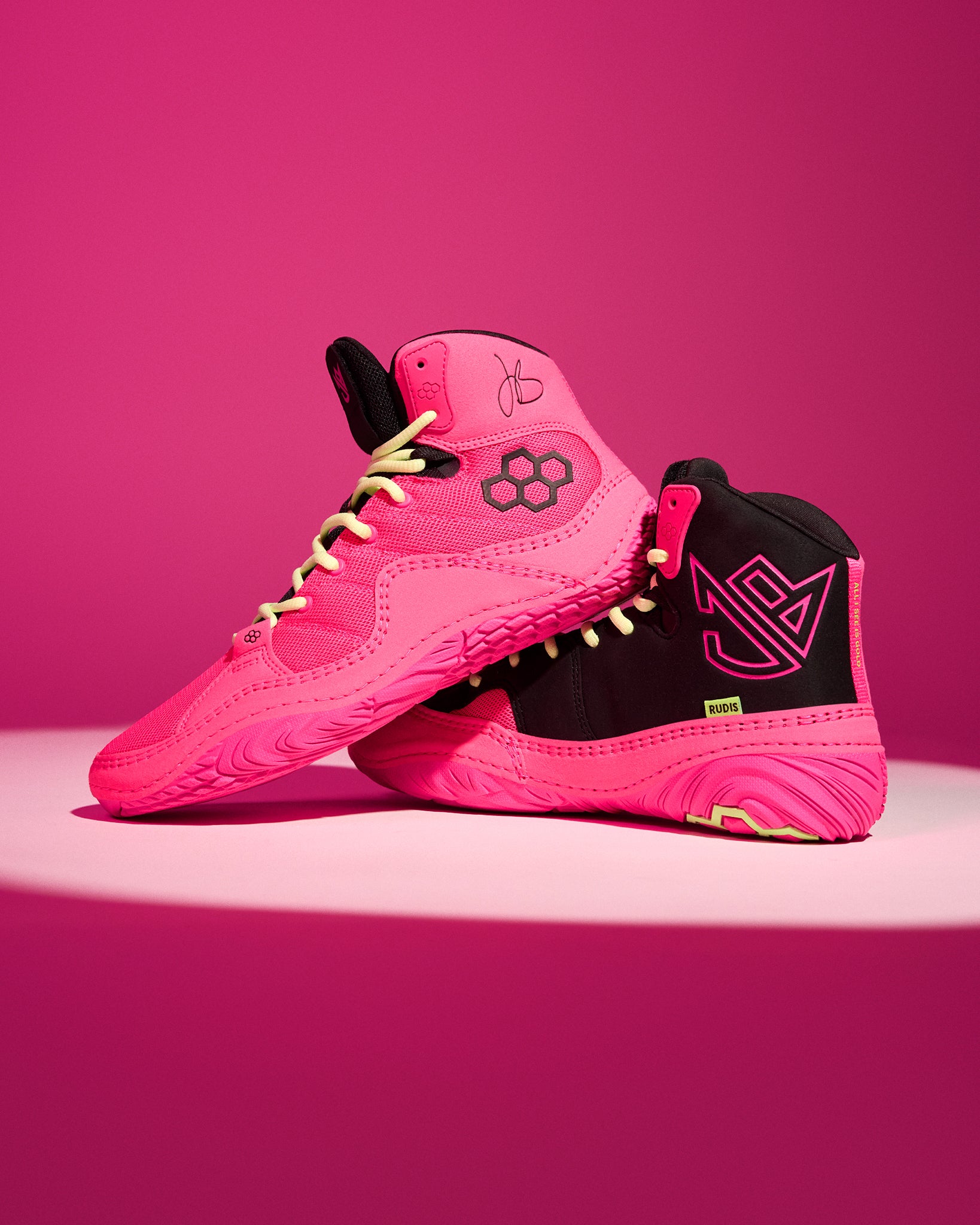 A pair of high-top wrestling shoe in vibrant pink and black, featuring a geometric flower logo on the side and a stylized 'Jordan Burroughs' logo on the heel, set against a solid pink background.