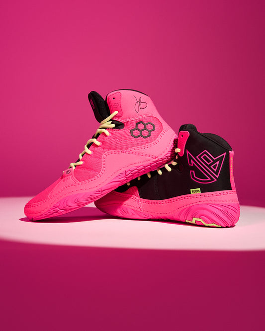A pair of high-top wrestling shoe in vibrant pink and black, featuring a geometric flower logo on the side and a stylized 'Jordan Burroughs' logo on the heel, set against a solid pink background.