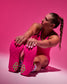 A woman in a pink athletic wrestling singlet sits on a pink studio floor, reaching toward her feet, with bright pink wrestling shoes featuring green geometric details visible. No visible brand text or logos are readable.