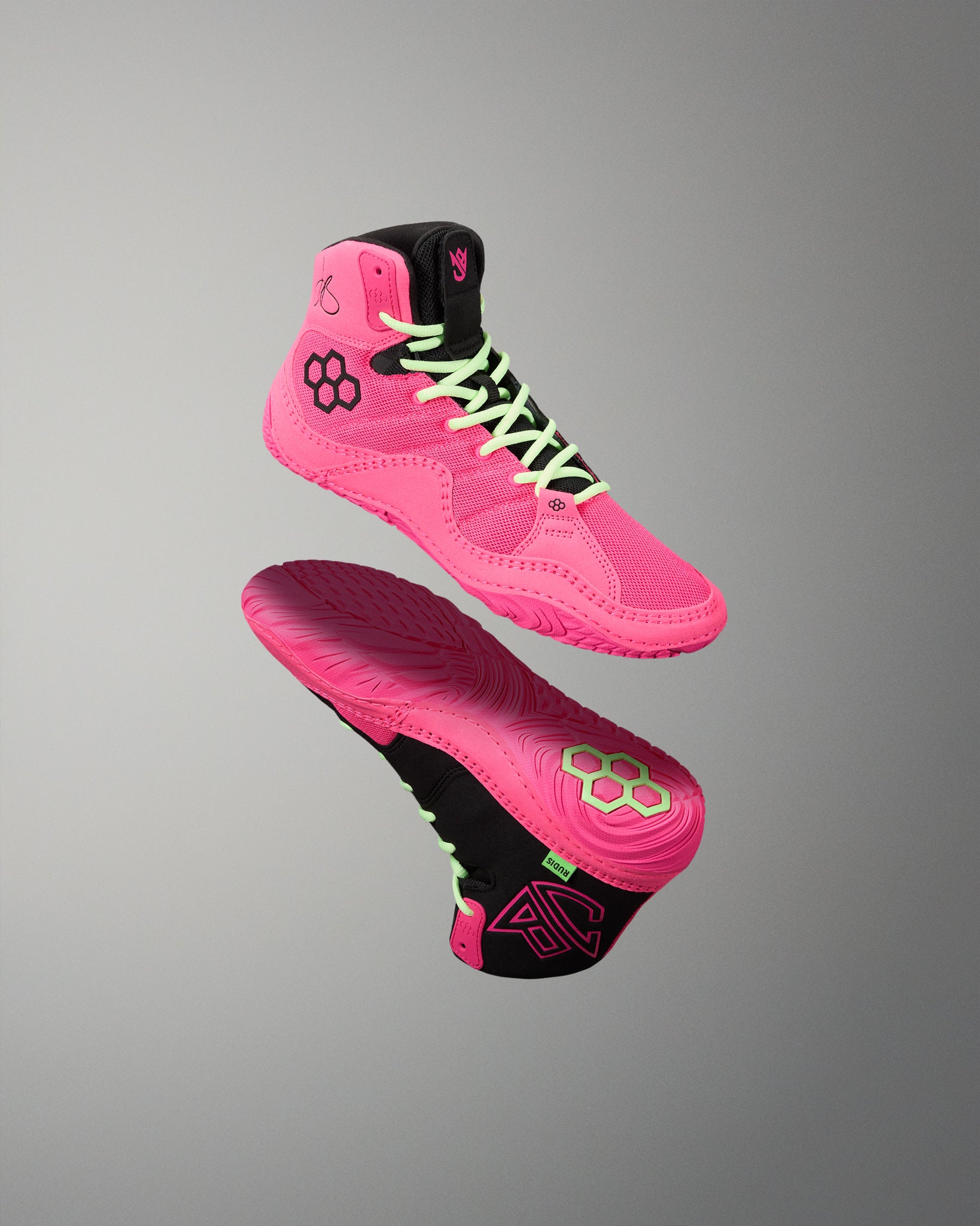 A pair of high-top athletic wrestling shoes in vibrant pink and black, shown floating against a neutral gray gradient background. One shoe is oriented to show the side profile with neon green laces, while the other shows the sole with a matching pink and black design.