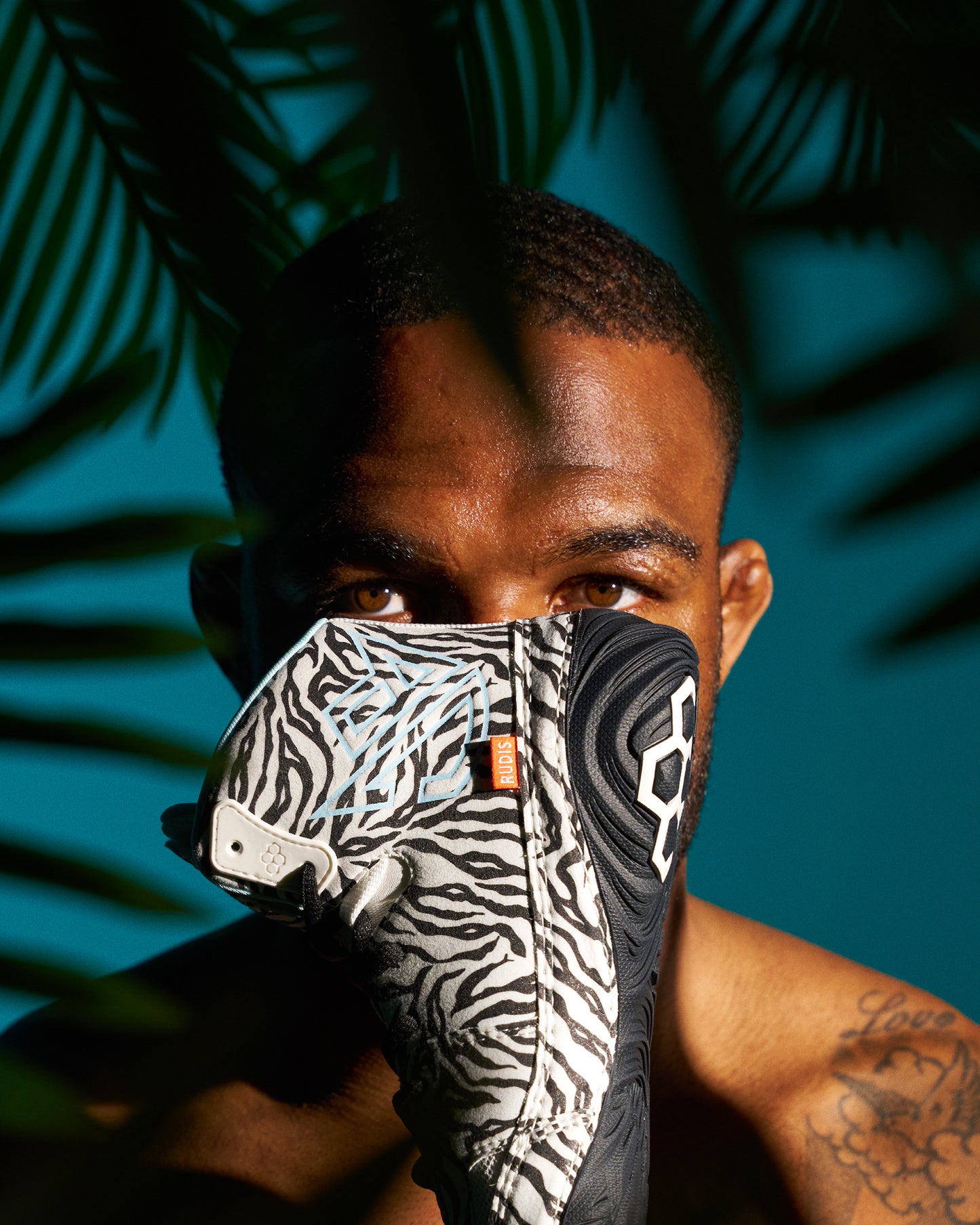 A close-up lifestyle shot of a man wearing a black and white zebra-patterned wrestling shoe in front of his mouth, set against a dark teal background with palm leaf shadows.