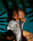 A close-up lifestyle shot of a man wearing a black and white zebra-patterned wrestling shoe in front of his mouth, set against a dark teal background with palm leaf shadows.