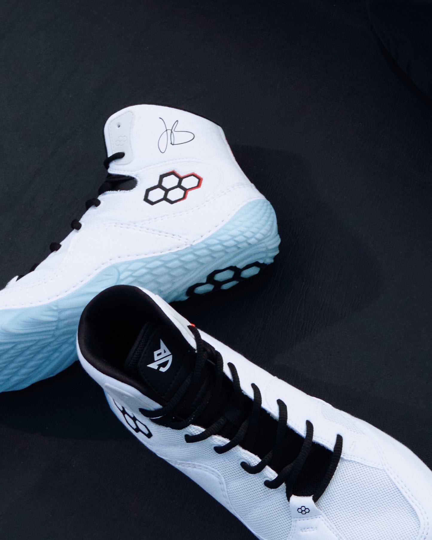 A pair of white high-top wrestling shoes with black laces and accents, featuring a light blue textured outsole and a red-and-black geometric logo on the side, displayed on a dark background.