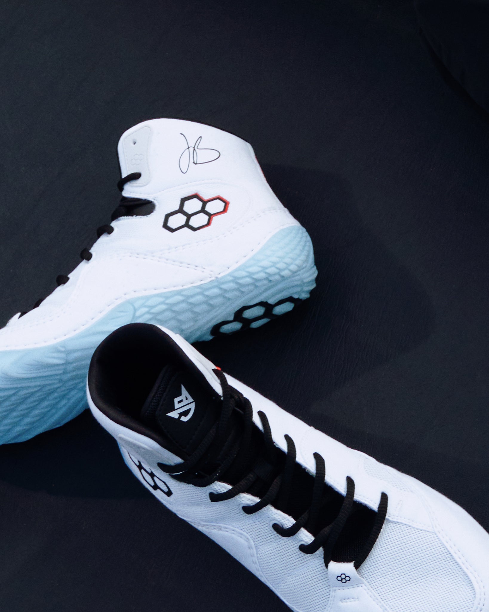 A pair of white high-top wrestling shoes with black laces and accents, featuring a light blue textured outsole and a red-and-black geometric logo on the side, displayed on a dark background.