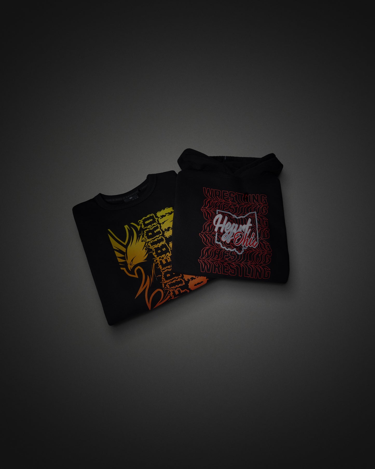 Two folded black Hoodie and Crewneck displayed on a dark gray gradient background. The left shirt features a yellow and orange graphic, and the right shirt features a red and white graphic with the words 'Heart of Ohio'.