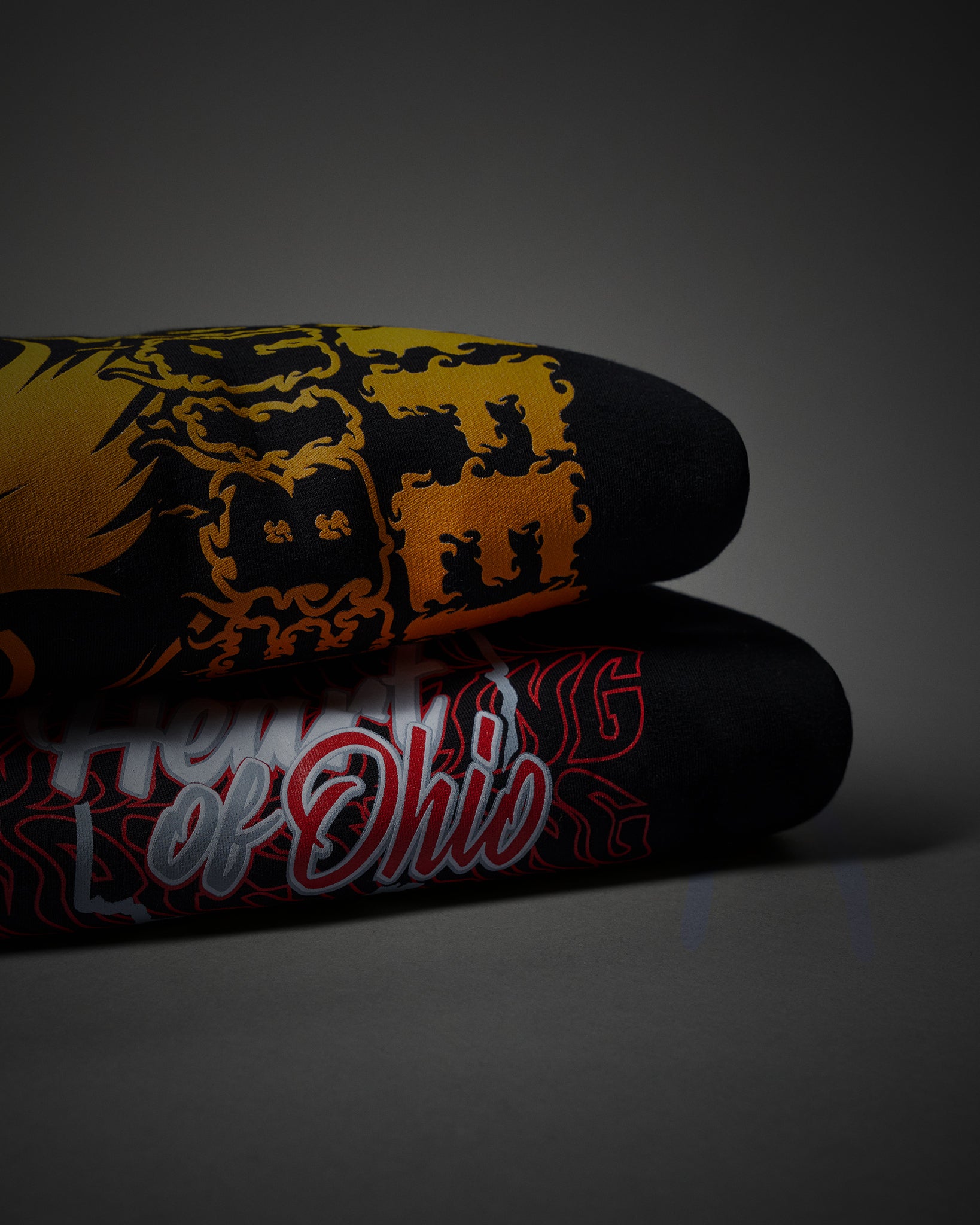 Two rolled black hoodie and crewneck stacked on a dark gray background. The top sleeve features a bold yellow and black graphic design, while the bottom sleeve displays 'HONOR OF OHIO' in stylized red, white, and black lettering.