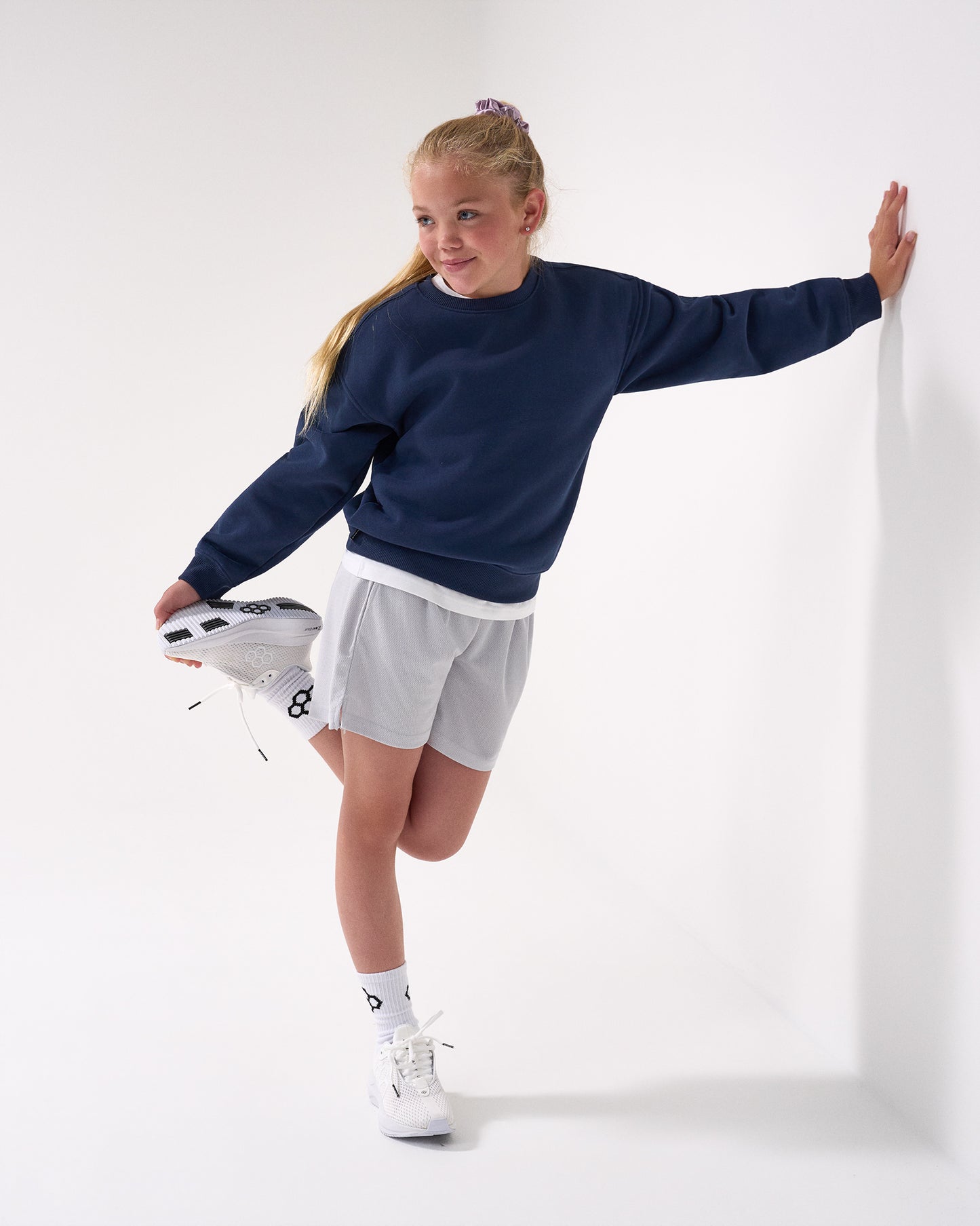 Young girl in navy sweater and gray shorts stretching against a white background