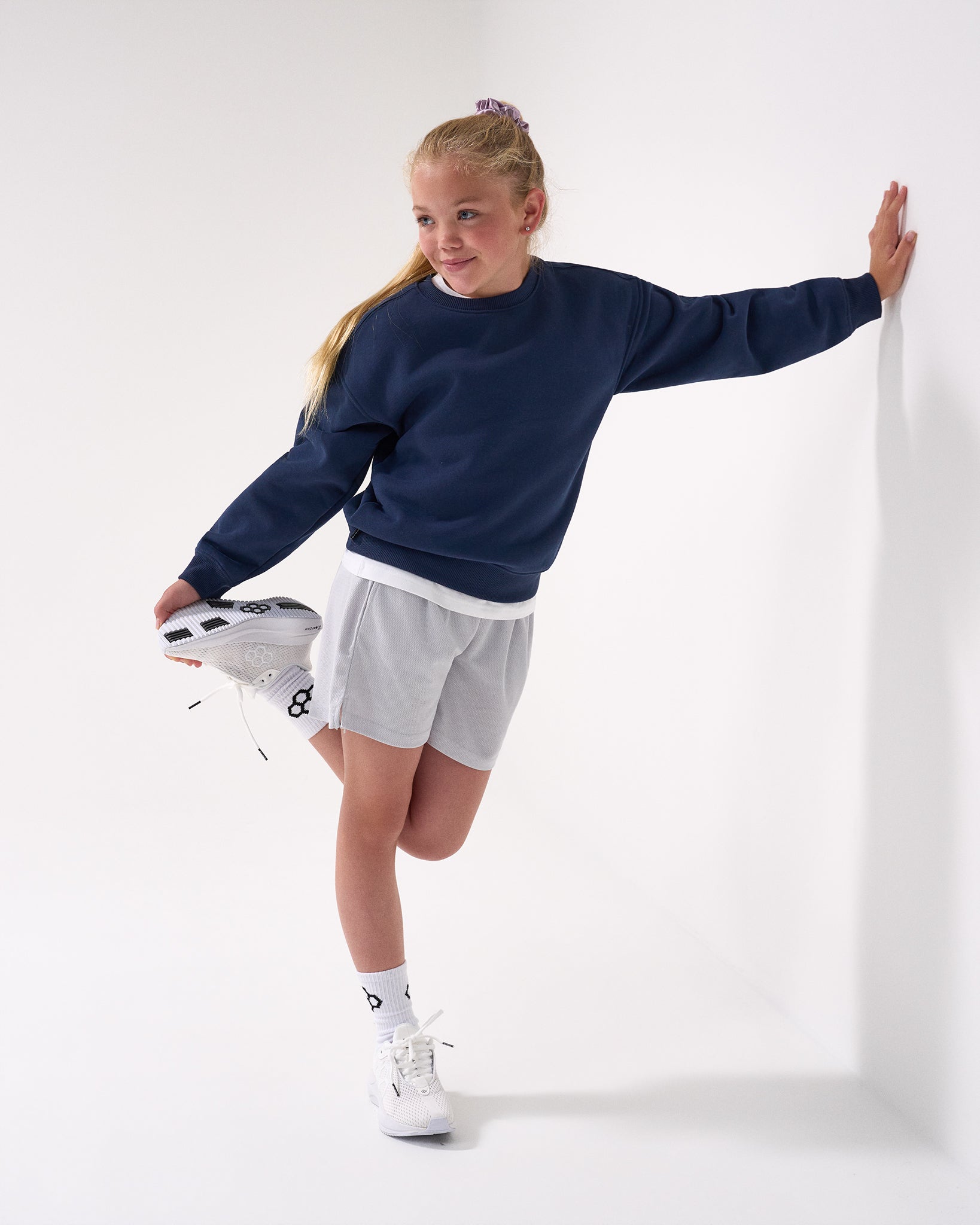 Young girl in navy sweater and gray shorts stretching against a white background
