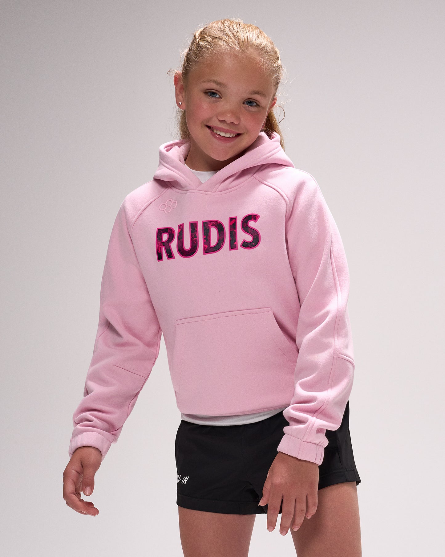 Young girl wearing a pink hoodie with 'RUDIS' text on a plain background