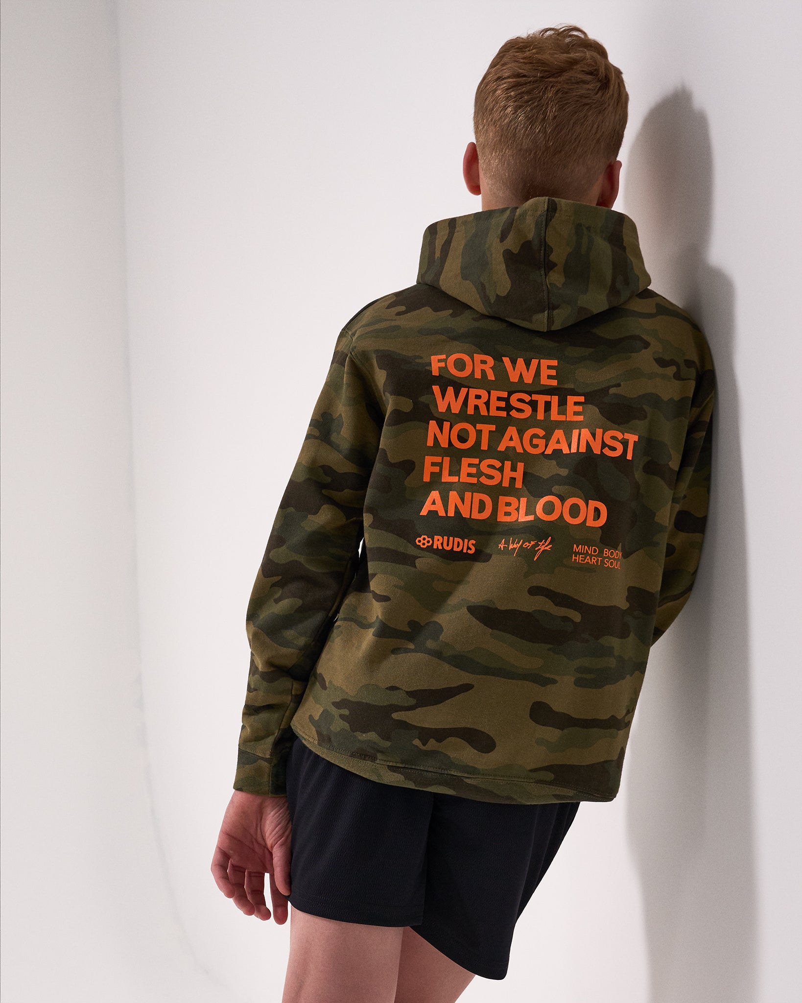 Person wearing a camouflage hoodie with 'For we wrestle not against flesh and blood' text on the back against a white wall