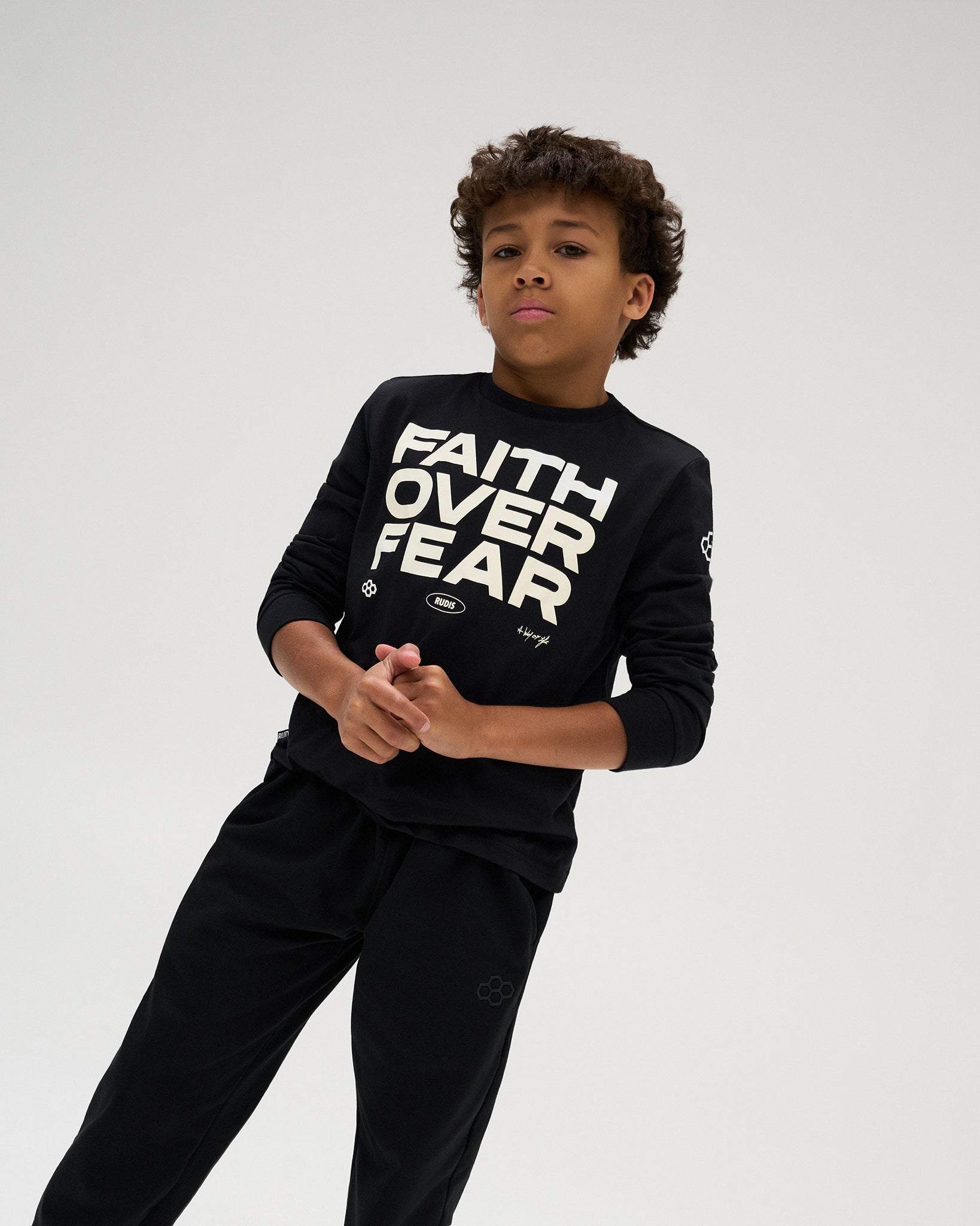 Youth athlete models the black RUDIS Faith Over Fear Youth Elite Super Soft Long Sleeve featuring bold “Faith Over Fear” graphics, paired with black athletic pants.