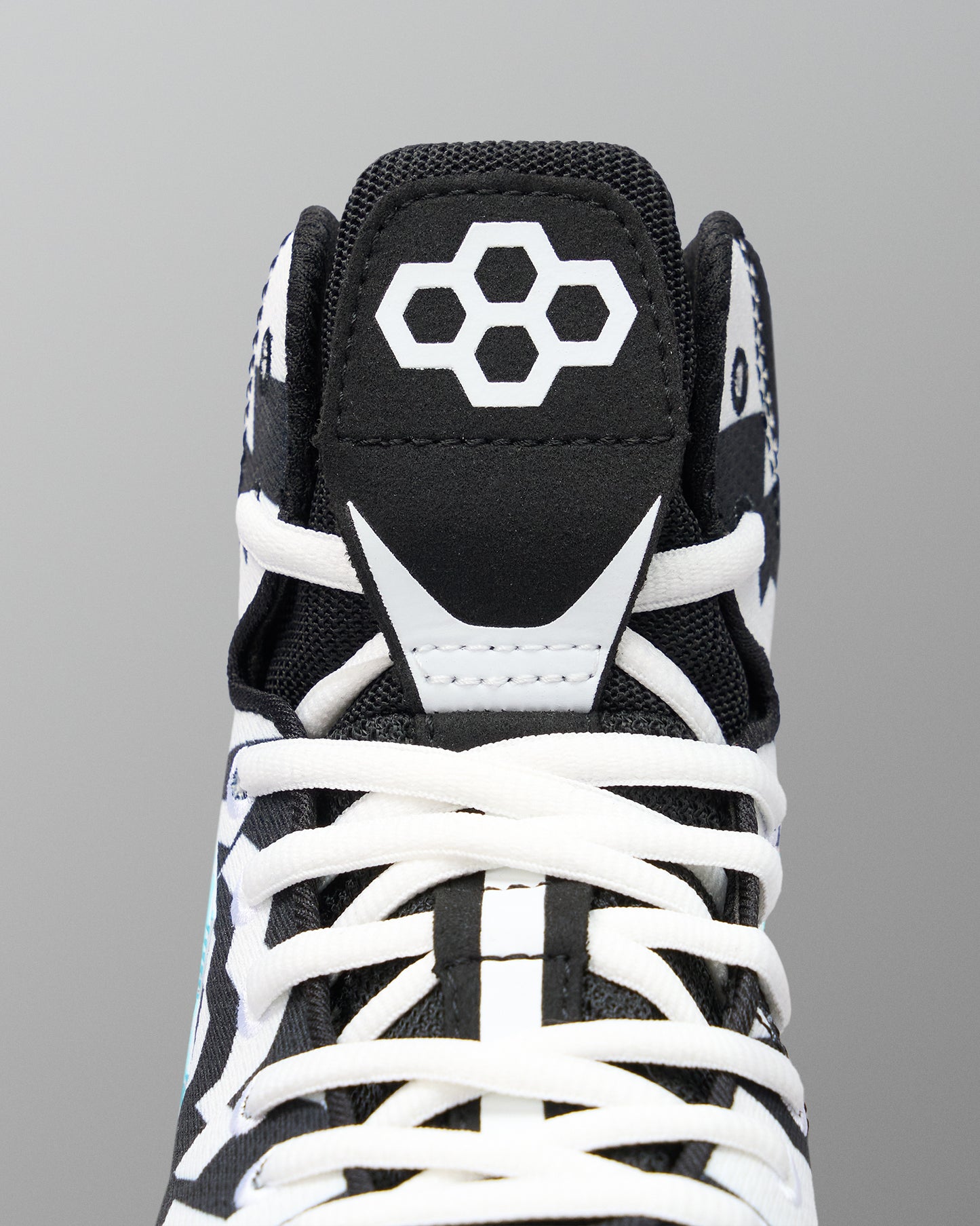 Close-up of a high-top wrestling shoe showing the black tongue with a white geometric logo, white laces, and white contrast overlays on a gray background.