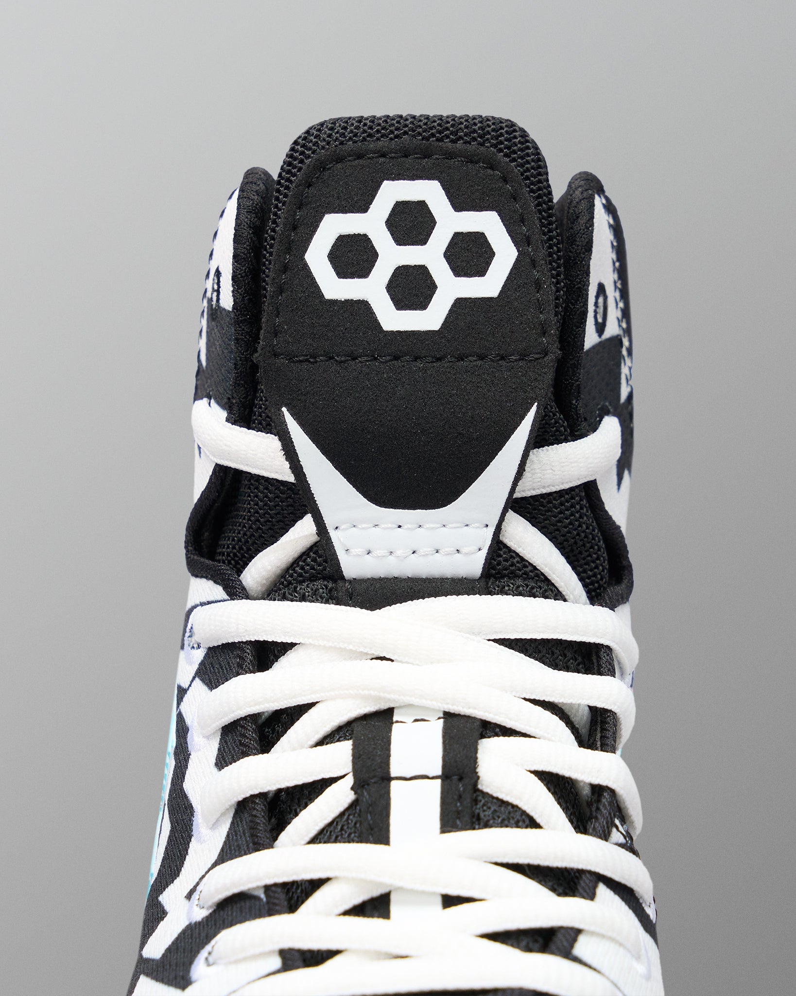 Close-up of a high-top wrestling shoe showing the black tongue with a white geometric logo, white laces, and white contrast overlays on a gray background.