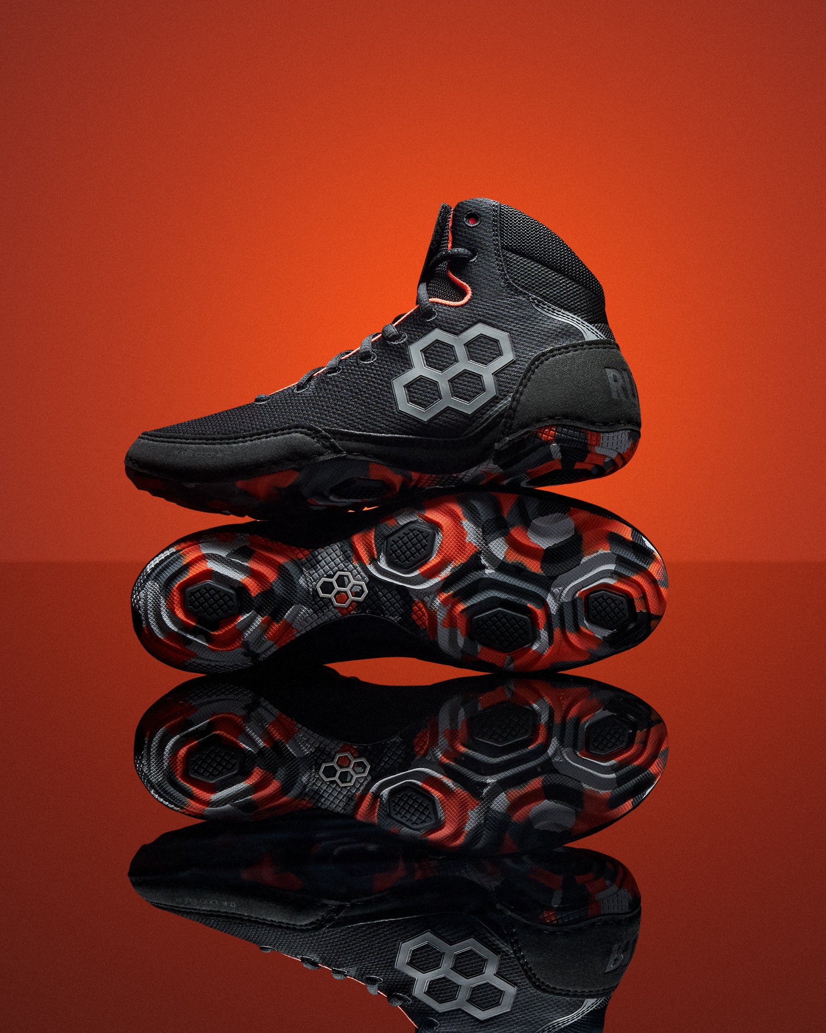 A pair of black high-top Wrestling Shoes with gray and red geometric hexagonal patterns, displayed on a reflective surface against a vibrant orange gradient background.